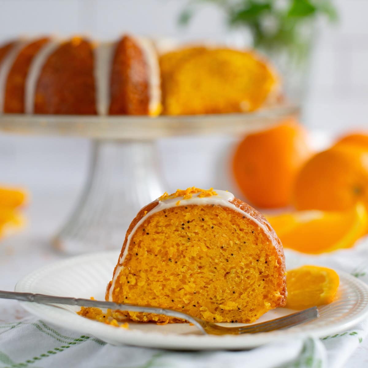 A close up image of orange poppy seed cake on a white plate with a fork in front. The whole cake is on a cake stand in the background.