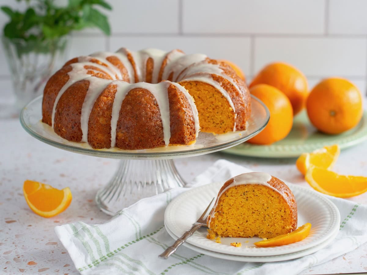 Orange poppy seed cake on a cake stand with a slice taken out. The slice is on a white plate in front with a fork on the side. A white and green linen is underneath with oranges in the background.