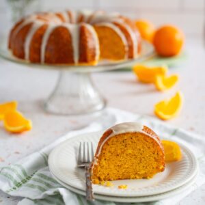 A close up image of a slice of orange poppy seed cake on a white plate with a fork. The whole cake is on a cake stand in the background.