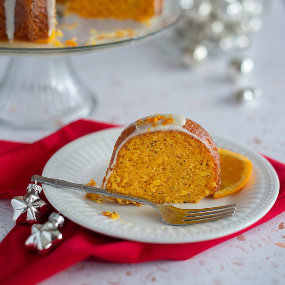 Slice of orange poppy seeds cake on white plate on red cloth next to silver Christmas ornaments.
