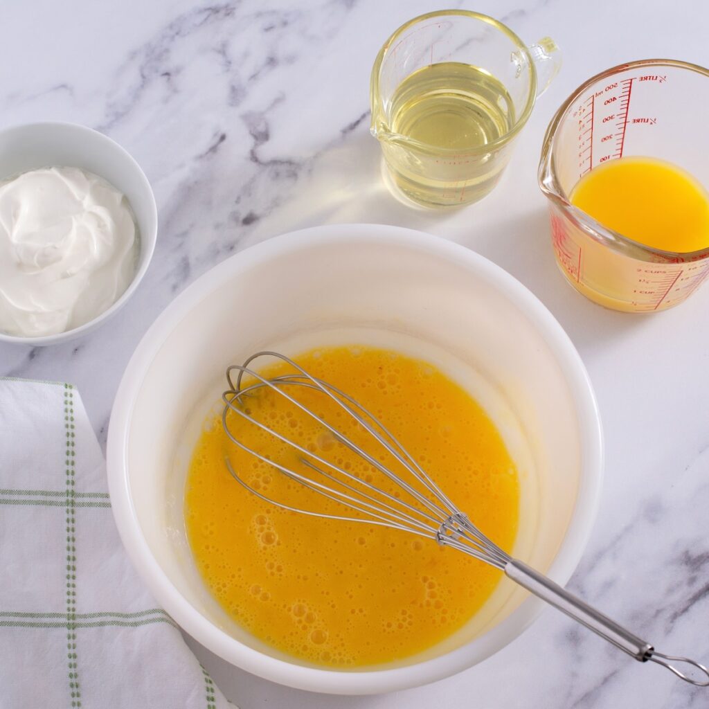 A white bowl with whisk eggs and a whisk resting on the side. Above is a white bowl of sour cream, a glass pitcher of oil, and a glass measuring cup of orange juice.