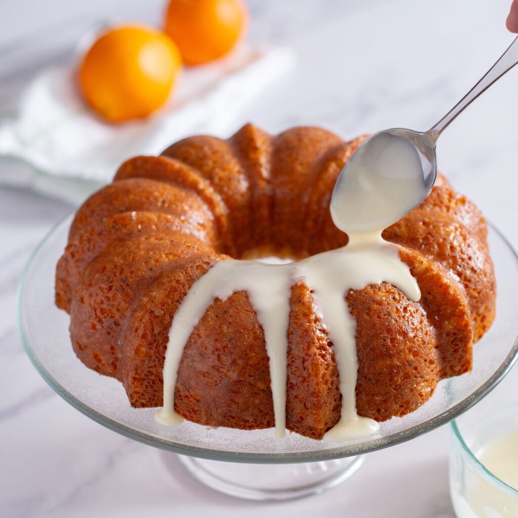 Orange poppy seed cake is on a glass cake stand. A spoon is drizzling over the orange glaze.