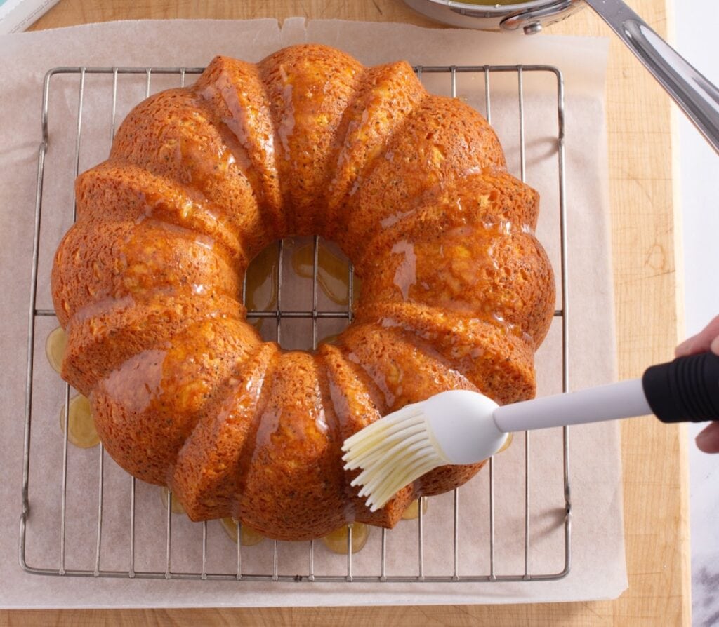 orange poppy seed bundt cake with a glaze being brushed on with white pastry 
brush