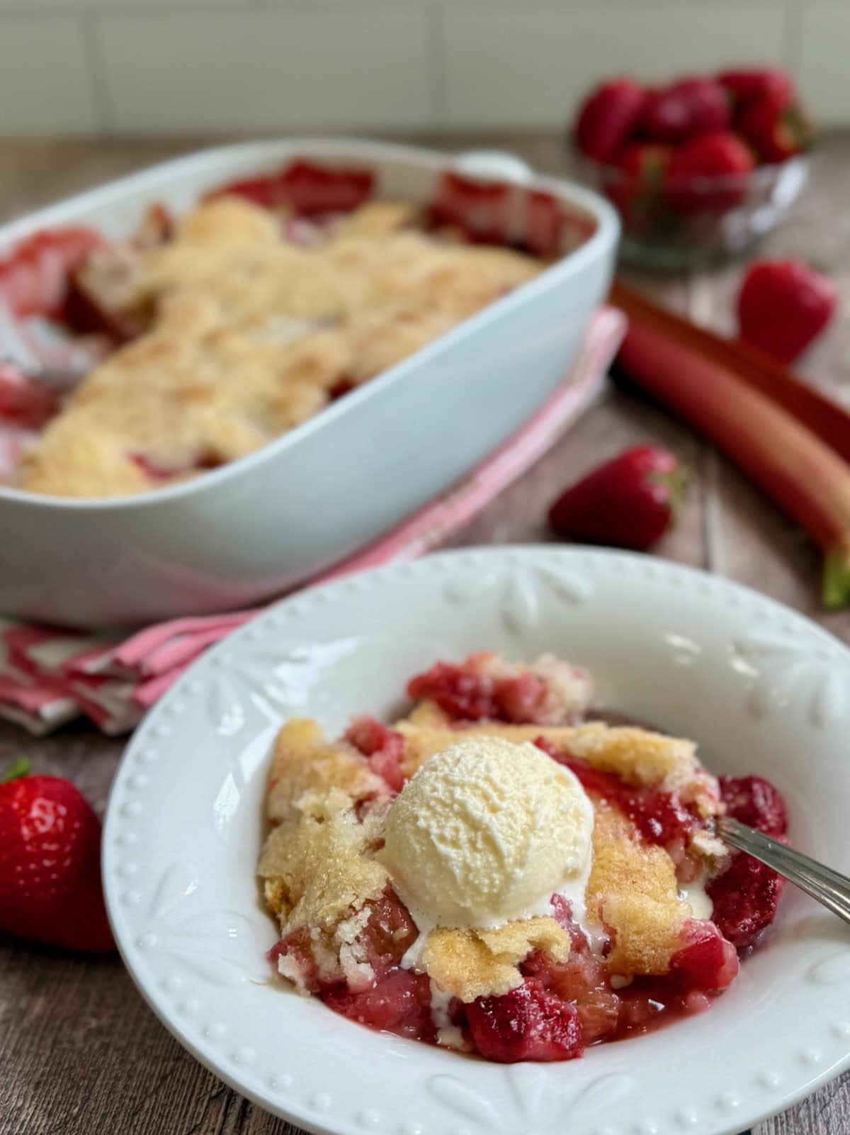 Bowl of strawberry rhubarb cobbler with ice cream, with the baking dish of cobbler in the background.