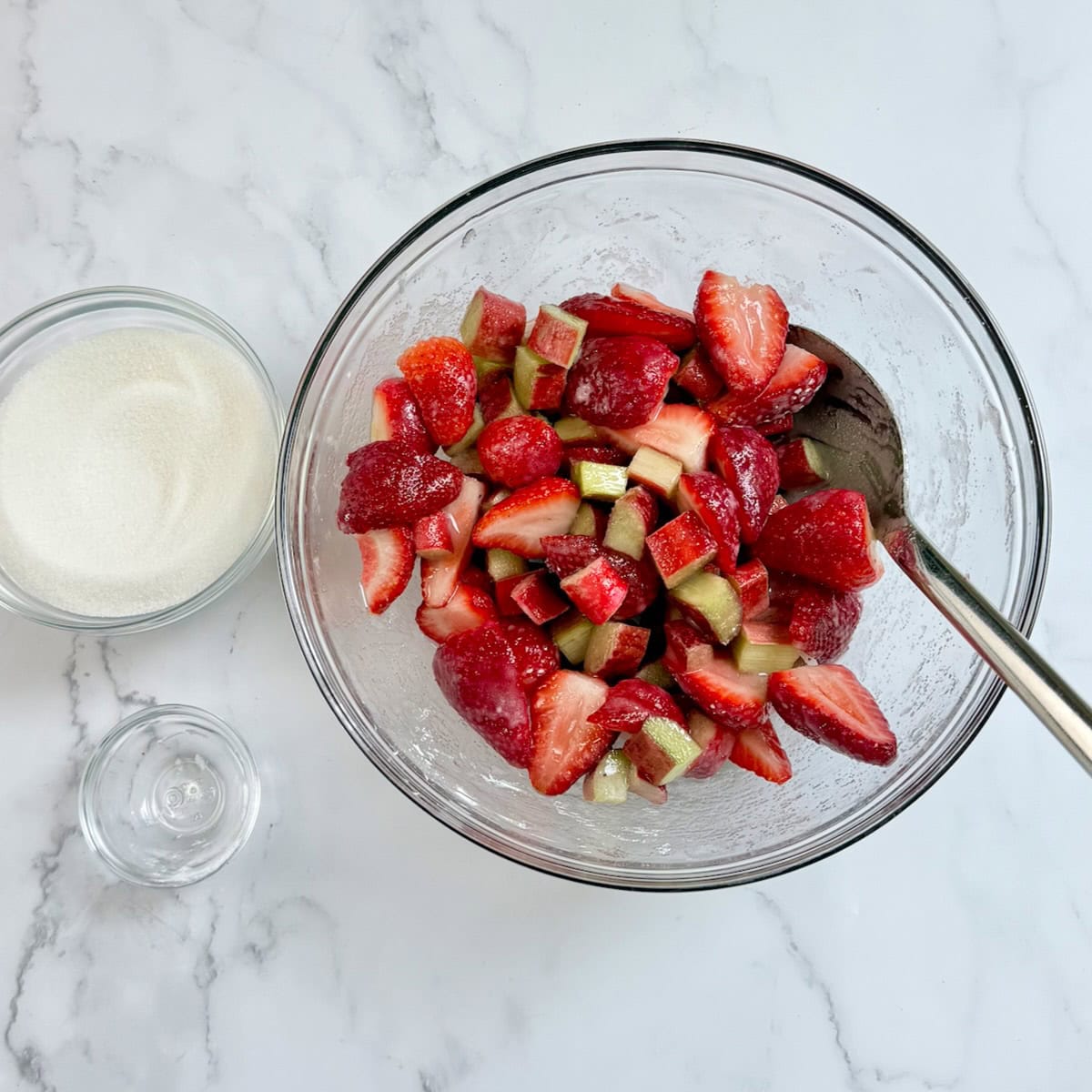 Fresh strawberries and rhubarb in a bowl with sugar next to it.