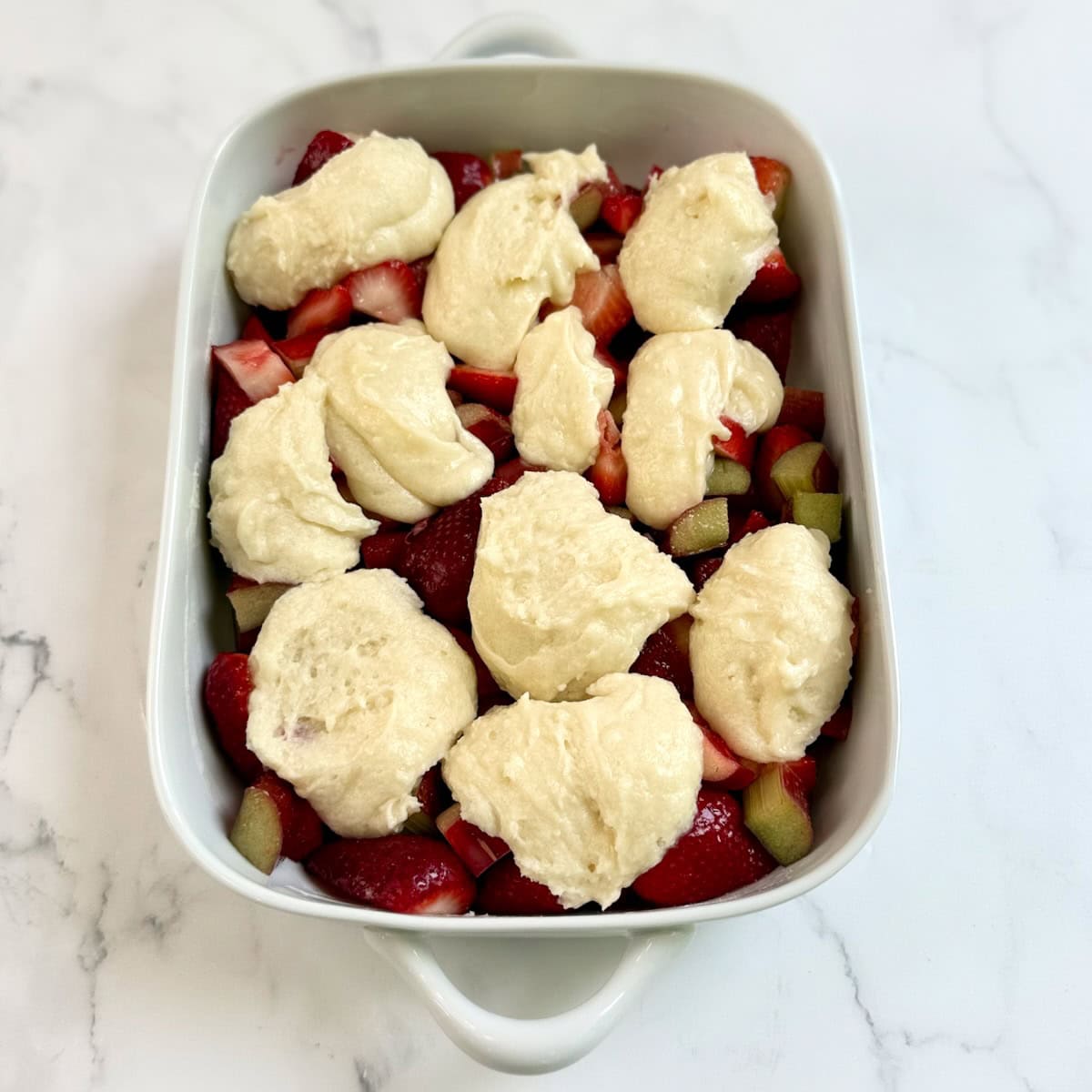 Fresh strawberries and rhubarb in the bottom of a baking dish with the cobbler dough dolloped on top.