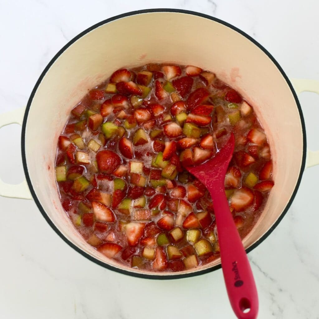 Fresh strawberry rhubarb jam cooking in a pot with a red spatula.
