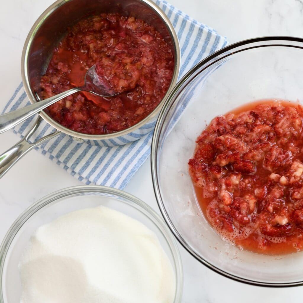 One bowl of mashed strawberries and a saucepan of cooked rhubarb next to a bowl of sugar.