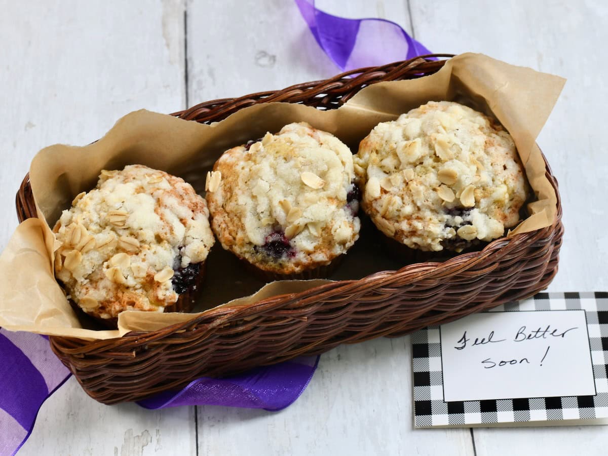 Three banana blackberry muffins in a brown wicker basket with a Feel Better Soon card next to it. 
