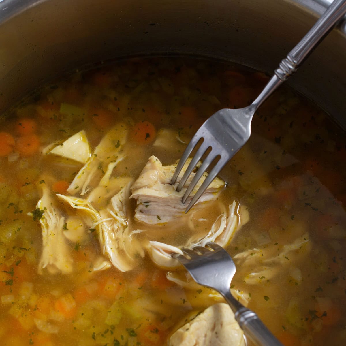 Two forks shredding chicken breast in a pot of soup.