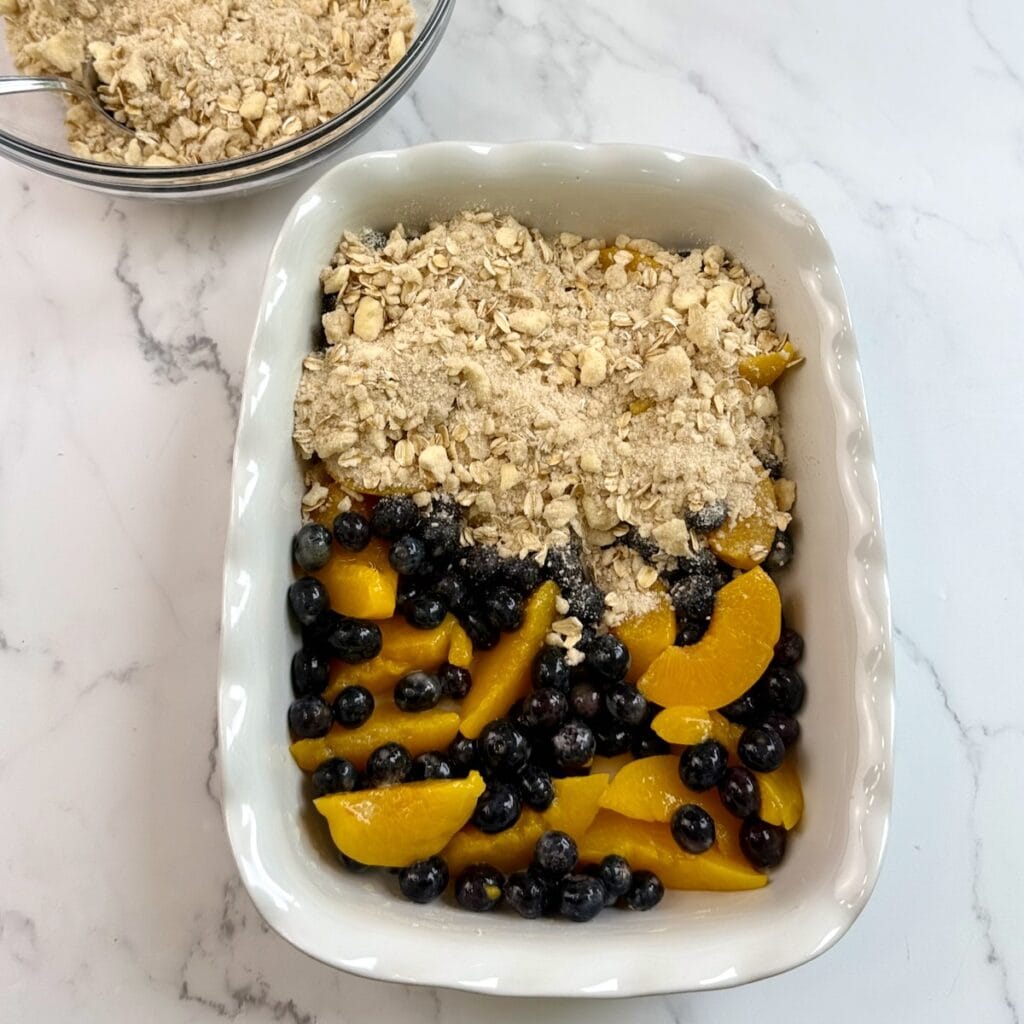 Assembling the peach blueberry cobbler in a white baking dish with half of the cobbler topping on top.