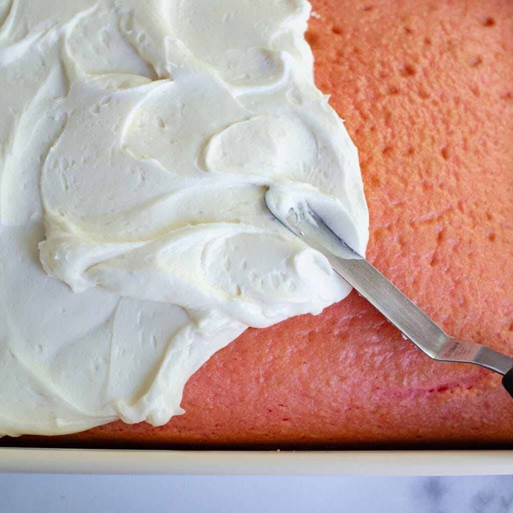 Using an offset spatula to spread cream cheese frosting on a strawberry cake.
