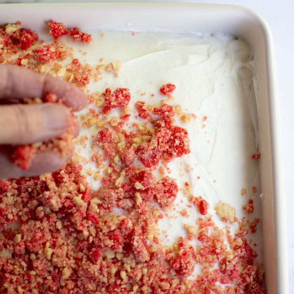 Hand sprinkling the strawberry crunch on top of the frosting layer of a cake.