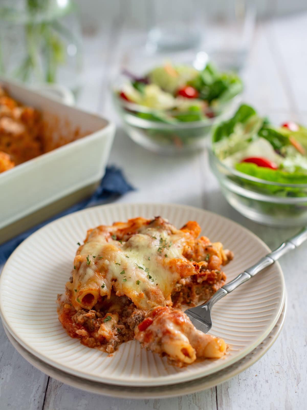 Baked ziti on a white serving plate with a fork scooping out a bite and salads in background.
