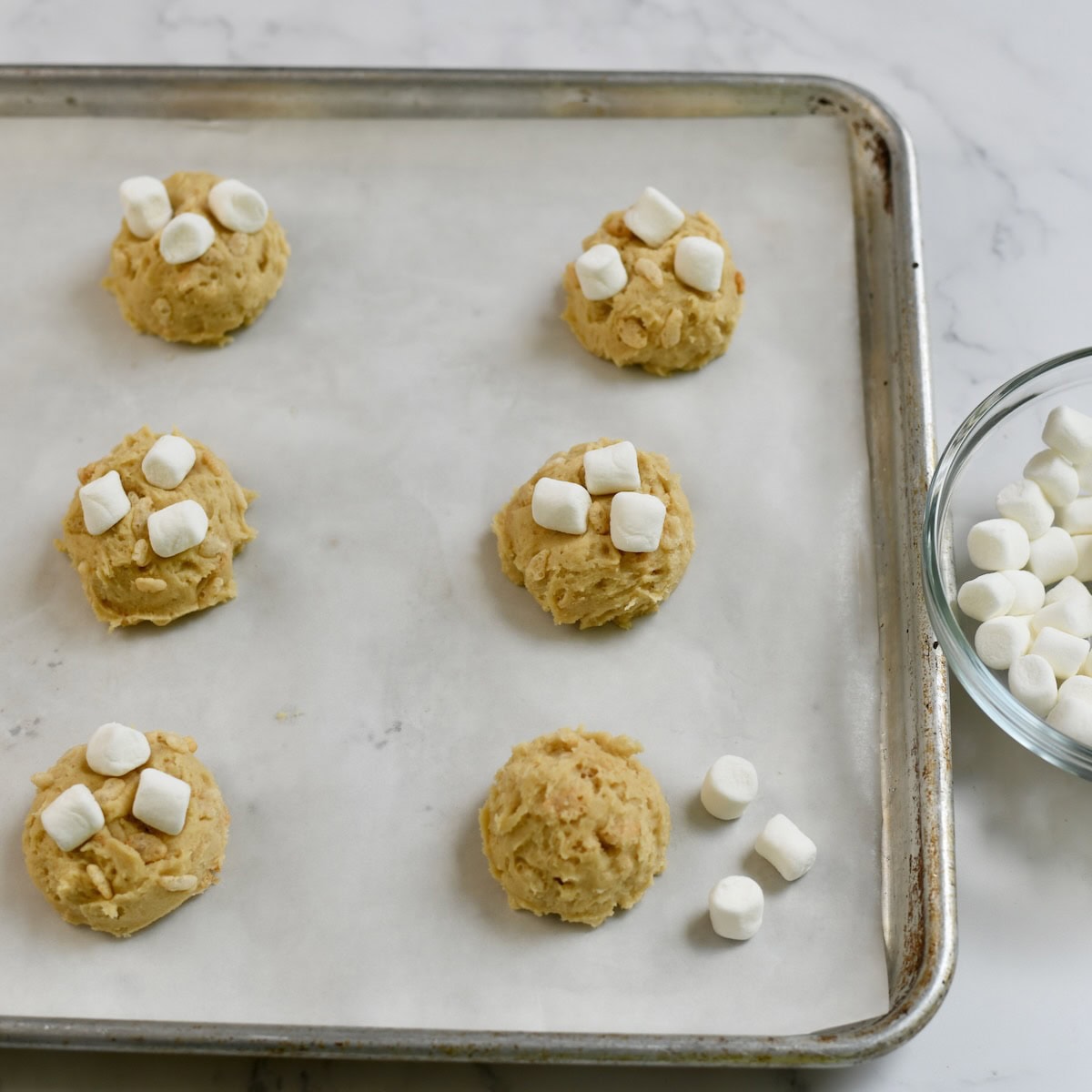 Rice krispie cookie dough balls on a sheet pan with some mini marshmallows on top.