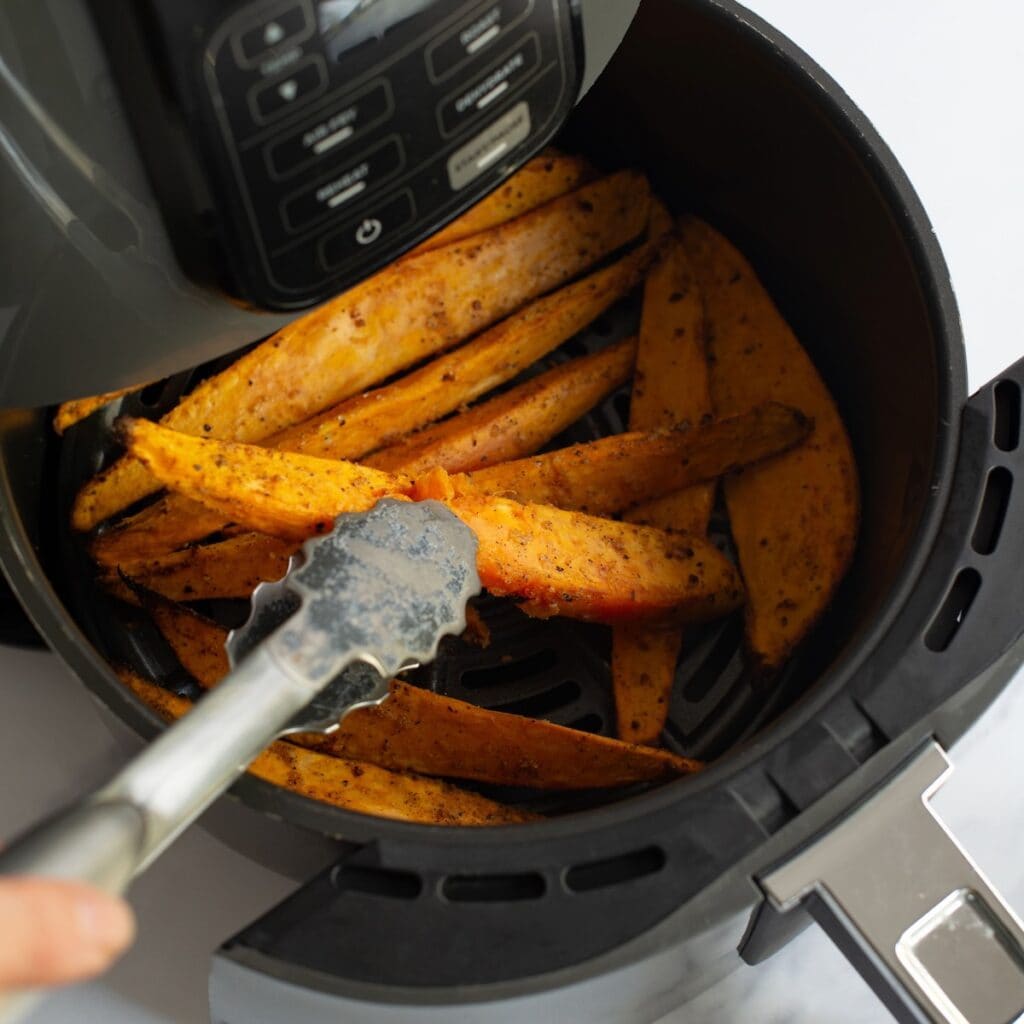 Tongs taking a sweet potato wedge from an air fryer basket.