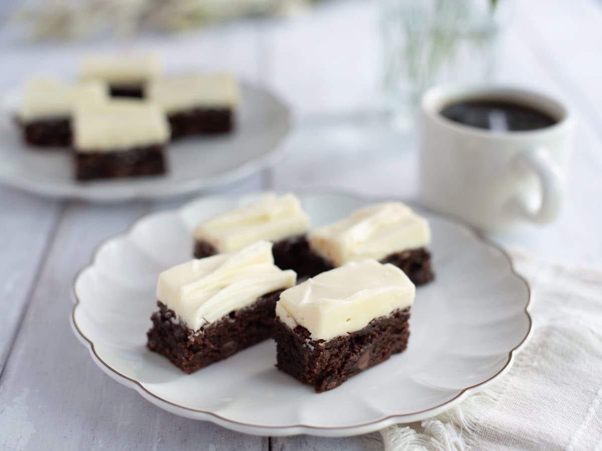 Four brownies with cream cheese frosting on a place next to a cup of coffee.