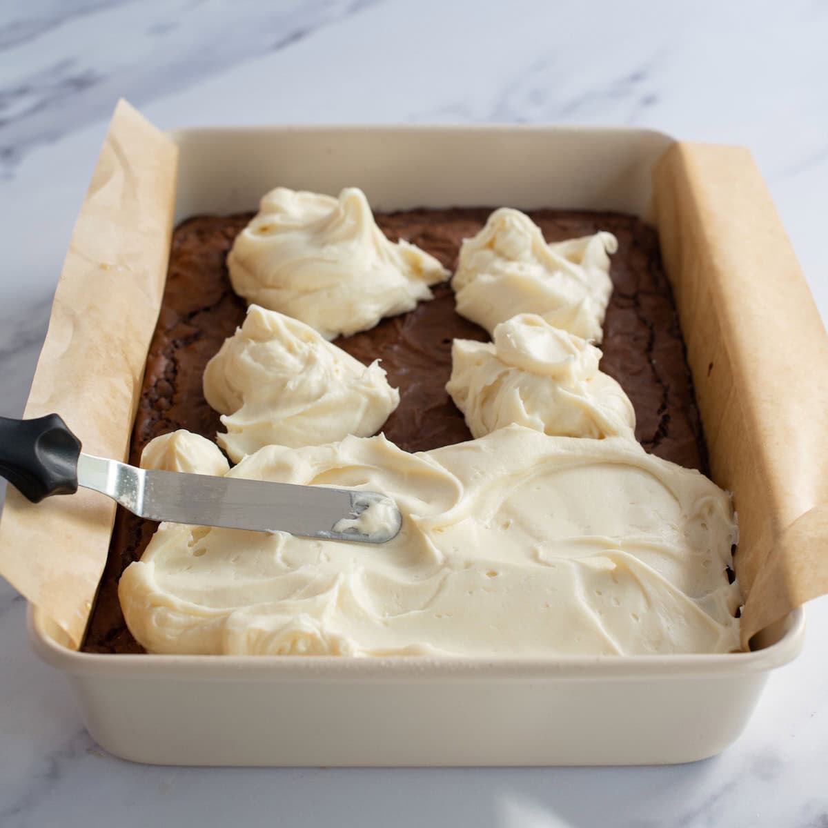 Spreading cream cheese frosting on the top of baked chocolate brownies.